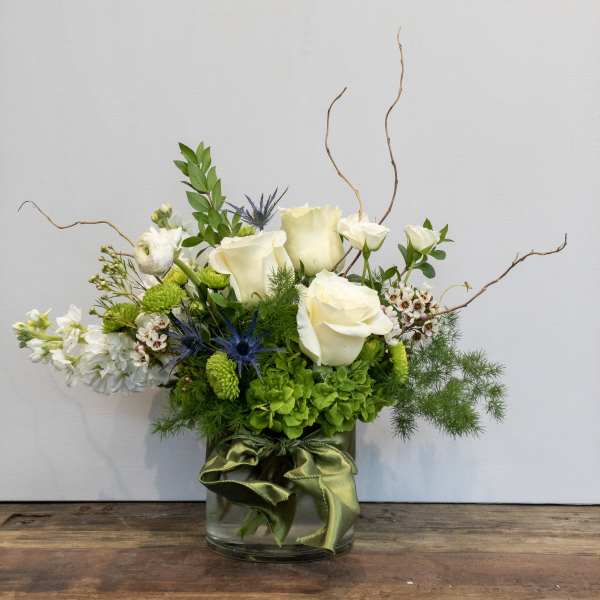 Low glass vase arrangement of white roses, white stock, green blooms, and blue thistle with a green ribbon.