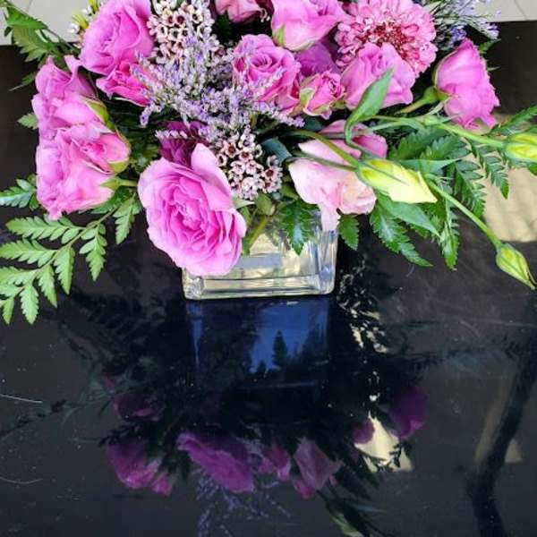 Low arrangement of pink roses and mixed purple flowers in a clear glass cube vase.