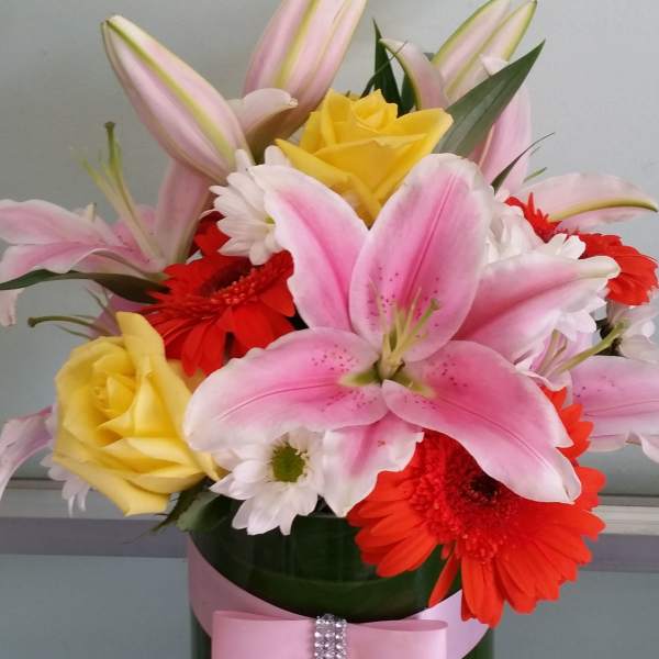 Pink lilies, yellow roses, and red gerbera daisies in a vase with a pink ribbon