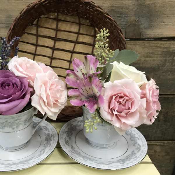 Two small floral arrangements in teacups on saucers with pink and purple roses