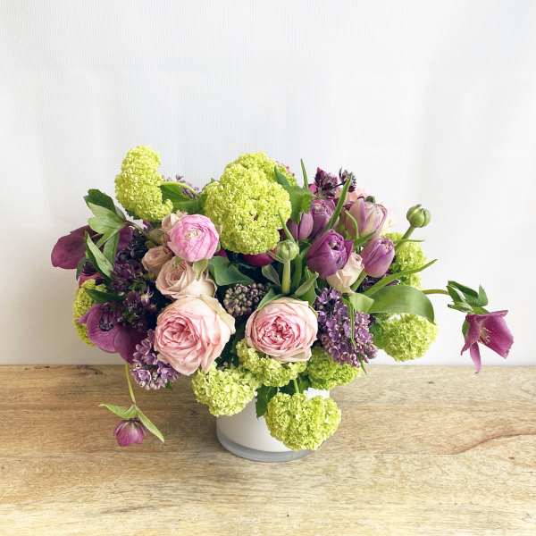 Low arrangement of pink roses, purple blooms, and green flower clusters in a white vase