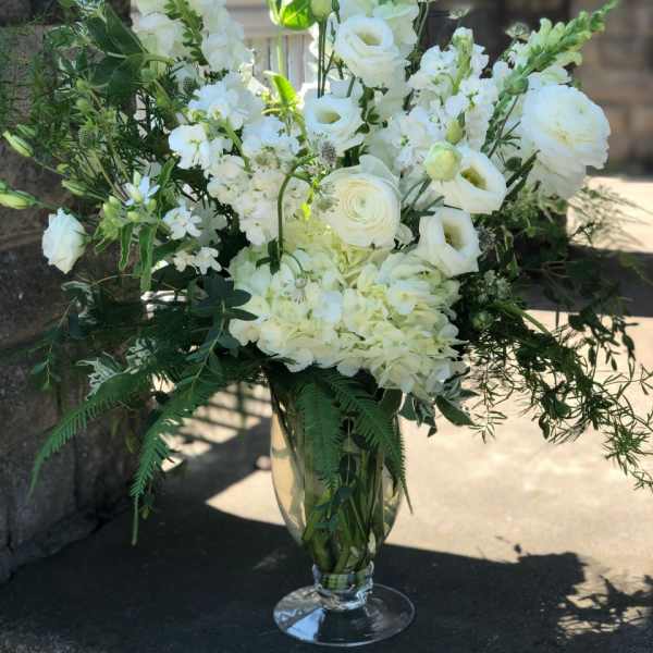 Tall all-white floral arrangement with roses, hydrangeas, and snapdragons in a clear glass pedestal vase