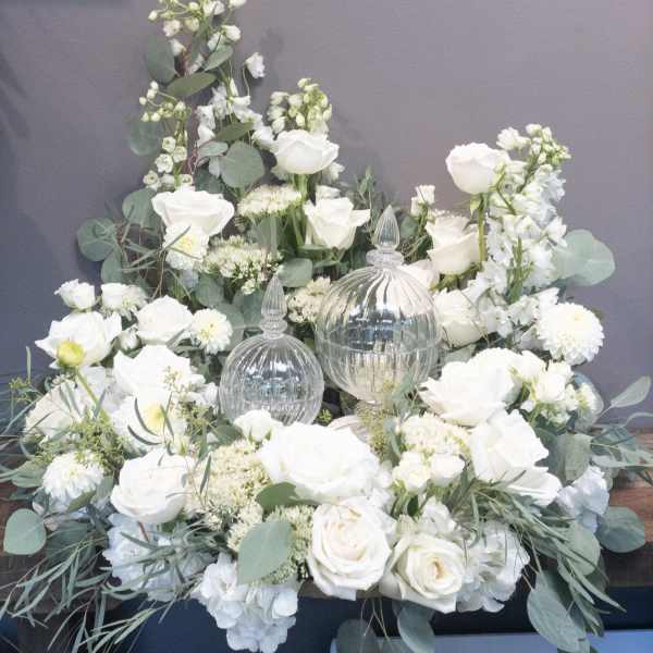 Low white flower arrangement with roses and mums surrounding two clear glass lidded jars.