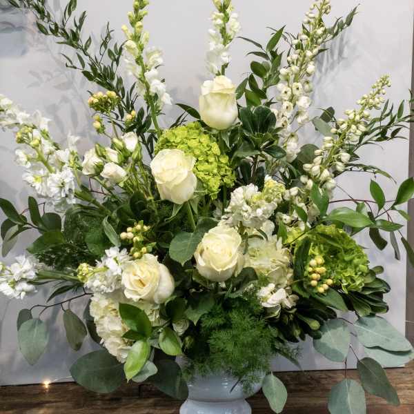 Tall white and green floral arrangement with roses, snapdragons, and hydrangeas in a white pedestal vase