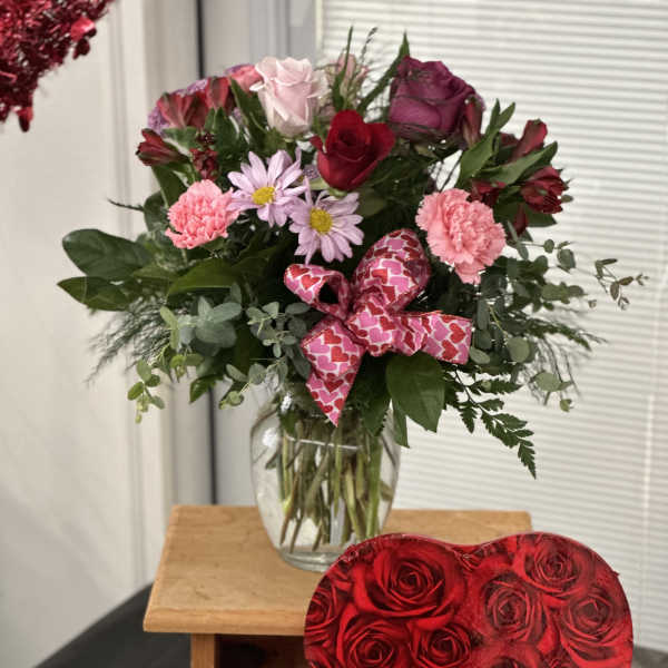 Mixed bouquet of roses and daisies in a glass vase with a heart ribbon