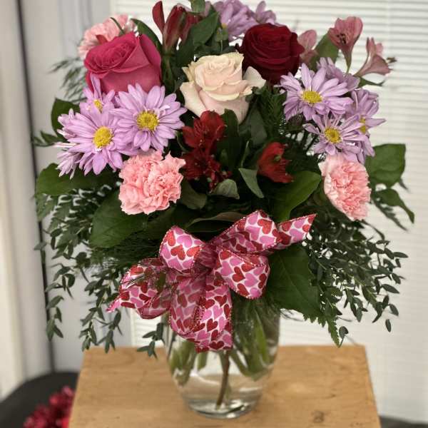 Mixed bouquet of roses, daisies, and carnations in a glass vase with a heart ribbon