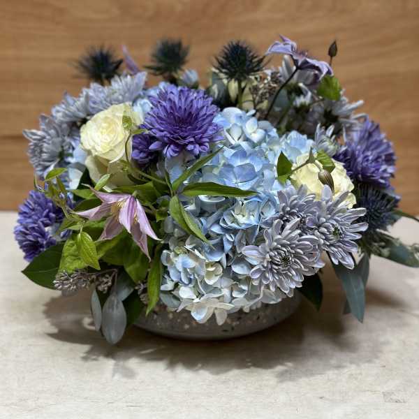 Low arrangement of blue hydrangeas, purple mums, and white roses in a shallow bowl