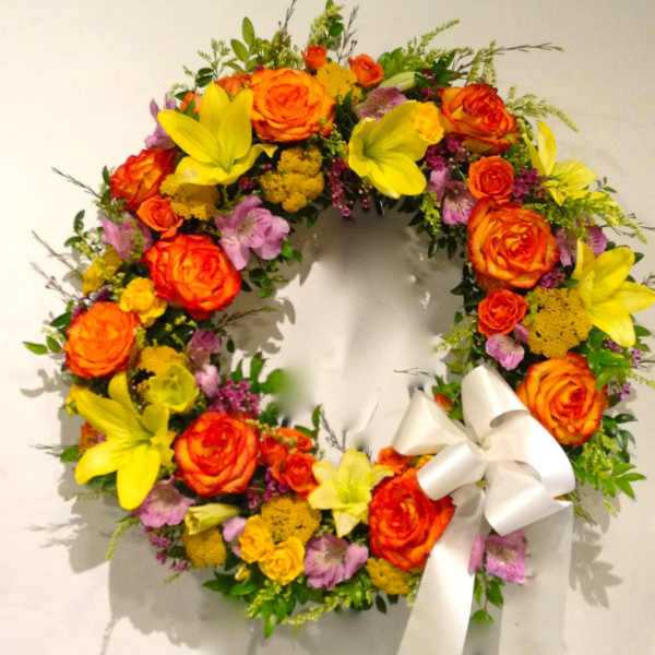 Circular floral wreath with orange roses, yellow lilies, and a white ribbon bow