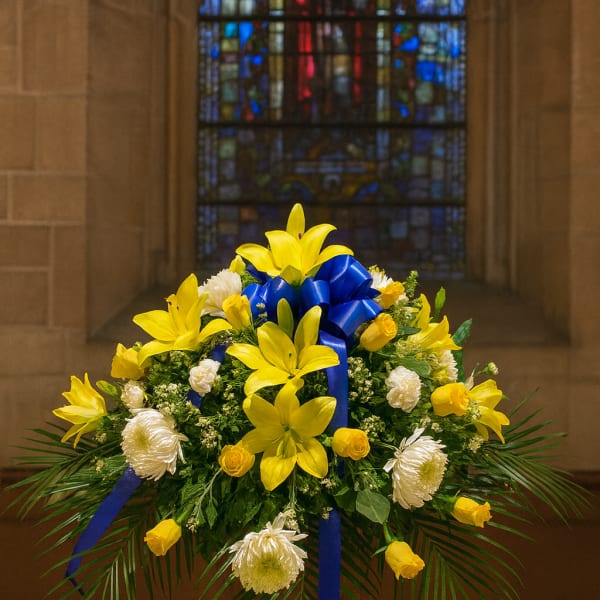 Yellow lilies and white flowers arranged with a blue ribbon on a pedestal.