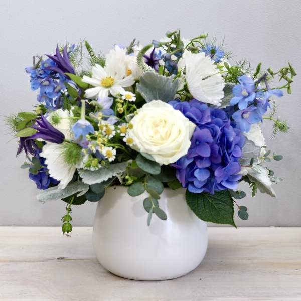 Short blue and white flower arrangement with hydrangeas and roses in a round white vase