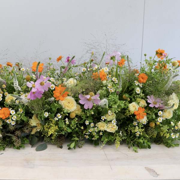 Long low arrangement of orange, yellow, pink, and white mixed flowers on a light wood surface