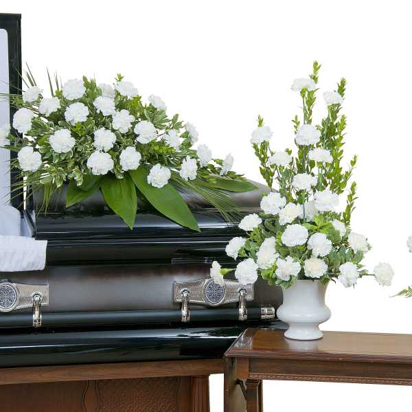 White carnation funeral casket spray with two matching vased arrangements beside a dark casket