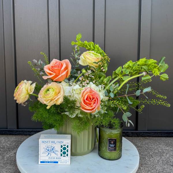 Peach and cream flower arrangement in a green vase with a candle and boxed soap on a marble tray