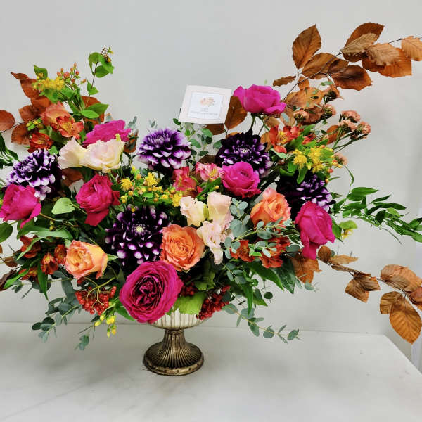 Wide arrangement of bright pink and orange roses with purple blooms and copper-toned branches in a pedestal vase