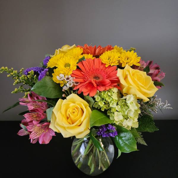 Mixed bouquet of yellow roses, gerbera daisies, and purple flowers in a glass vase