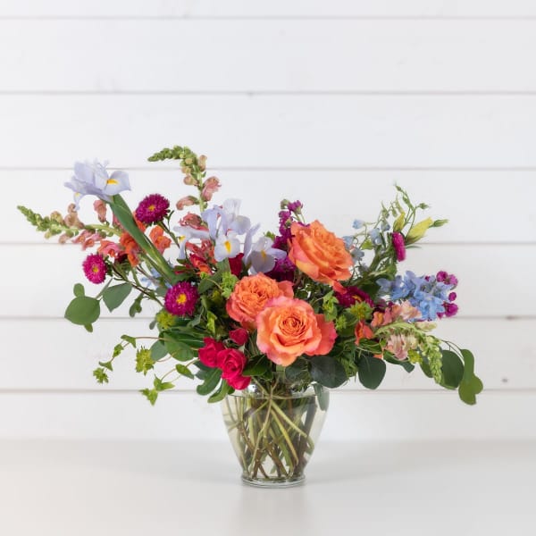 Mixed bouquet of orange, pink, and blue flowers in a clear glass vase