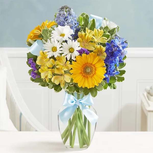 Mixed bouquet of daisies, gerberas, and blue flowers in a glass vase with a ribbon