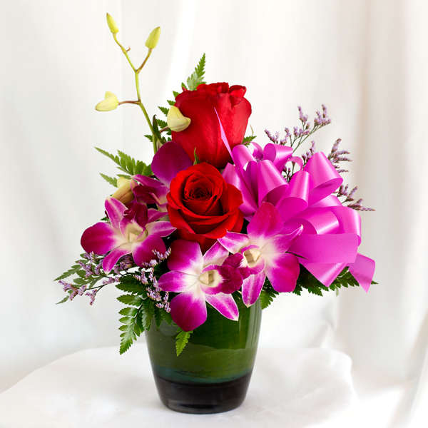 Red roses and purple orchids in a green glass vase with a pink ribbon