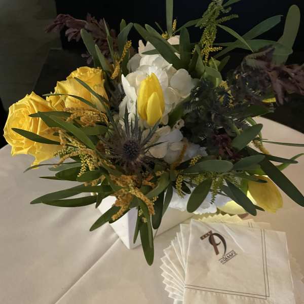 Yellow roses and white flowers arranged in a white box with napkins beside it.