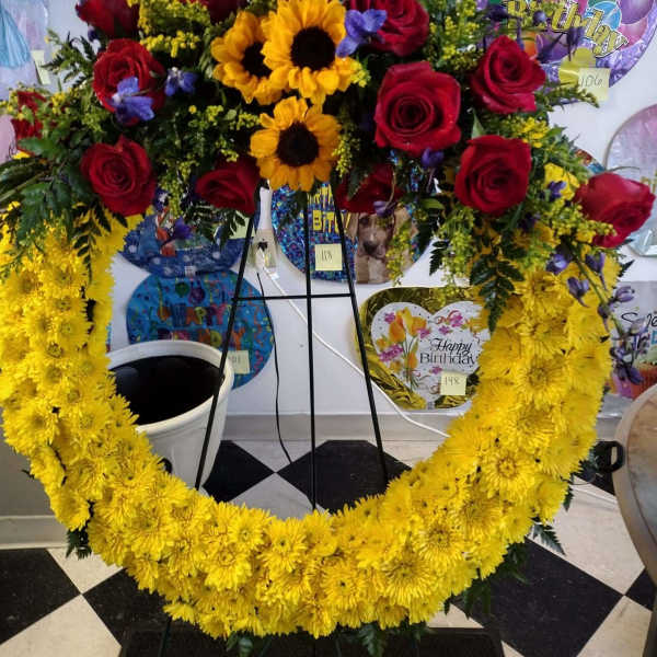 Heart-shaped floral wreath with red roses, yellow mums, and sunflowers on a stand