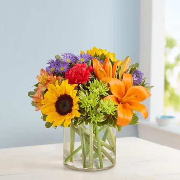 Mixed bouquet of sunflowers, lilies, and purple flowers in a clear glass vase