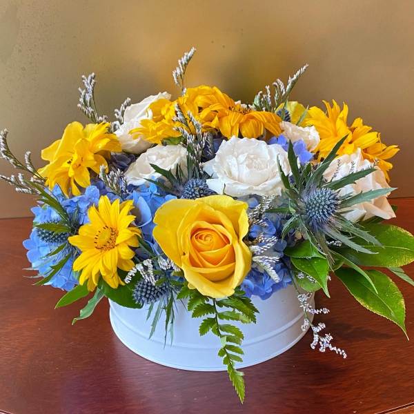 Yellow roses and daisies arranged with blue hydrangeas in a white container