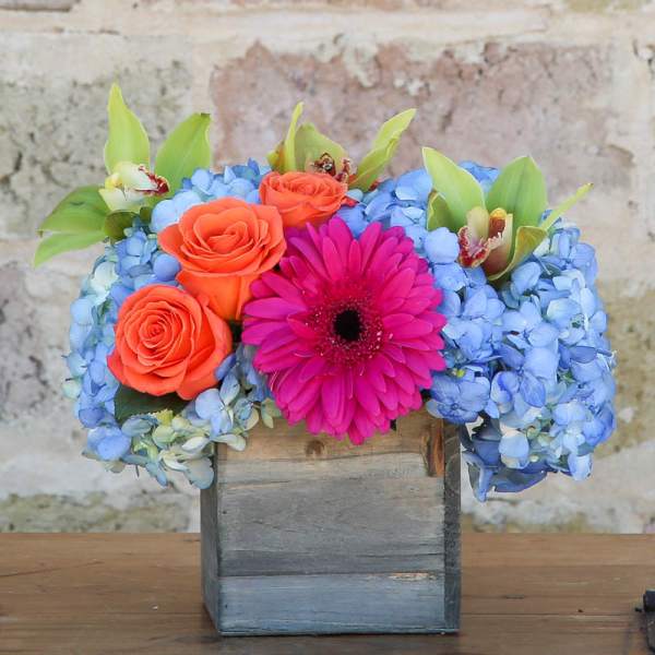 Bright bouquet of orange roses, blue hydrangeas, and a pink gerbera in a wooden box