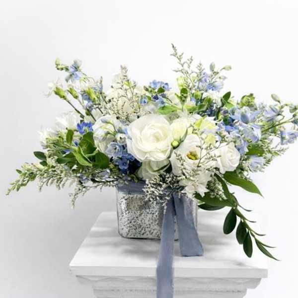 Low white and blue flower arrangement with roses and delphinium in a silver cube vase with blue ribbon