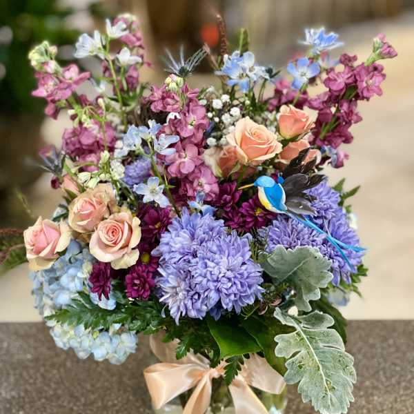 Mixed bouquet of pastel roses, purple blooms, and blue flowers in a glass vase