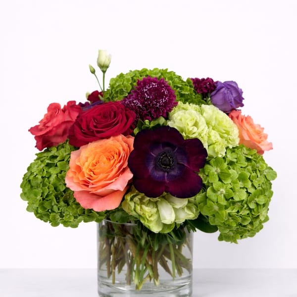 Colorful bouquet of roses, hydrangeas, and a dark anemone in a glass vase