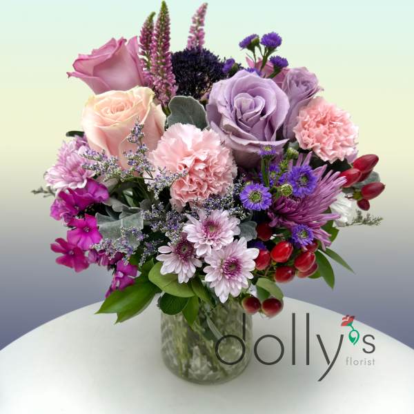 Mixed bouquet of pink and lavender roses, carnations, and purple blooms in a clear glass vase.