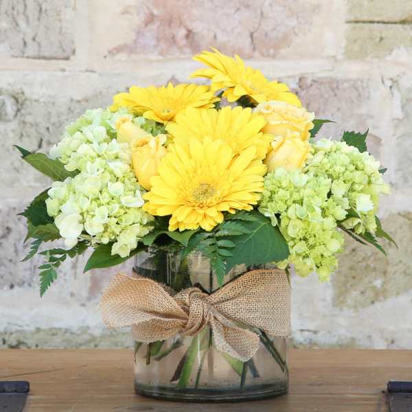 Yellow daisies and pale green hydrangeas in a glass vase with a burlap bow