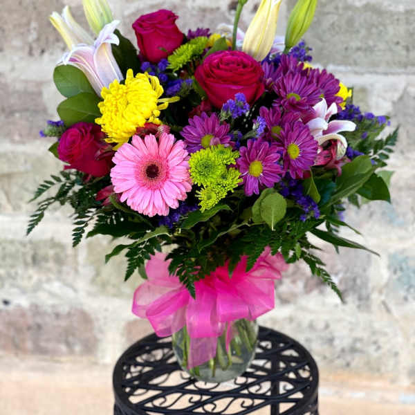 Mixed bouquet of pink roses, lilies, daisies, and mums in a glass vase with a bright pink bow