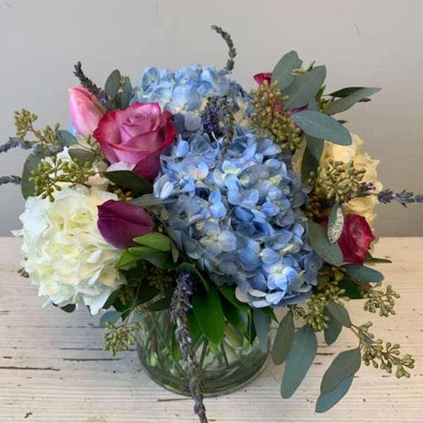 Low glass vase with blue hydrangeas, white blooms, and pink roses in a rounded arrangement.