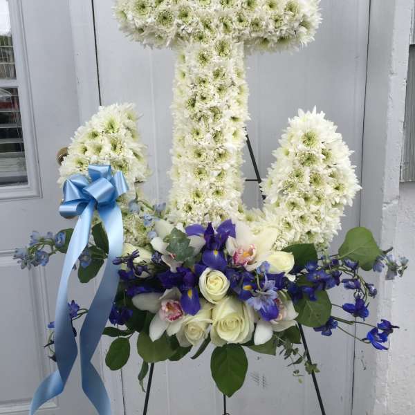 White floral cross on an easel with blue ribbon and purple flowers at the base