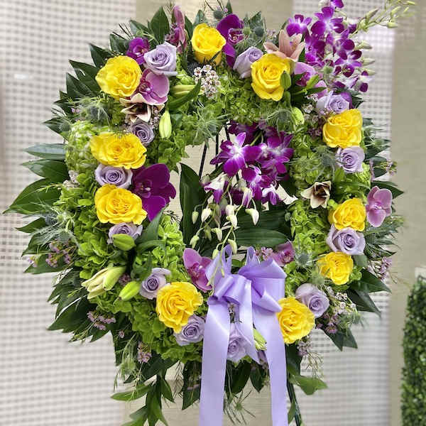 Standing wreath of yellow roses, lavender roses, and purple orchids with a lavender bow on an easel.