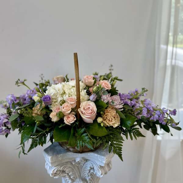 Pastel pink roses, white hydrangea, and lavender blooms arranged in a handled basket.
