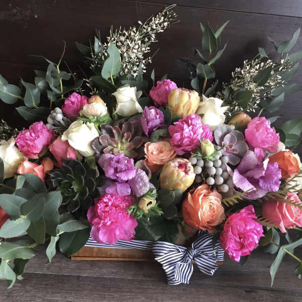 Low rectangular arrangement of pink peonies, mixed roses, succulents and protea in a wooden box with a striped bow
