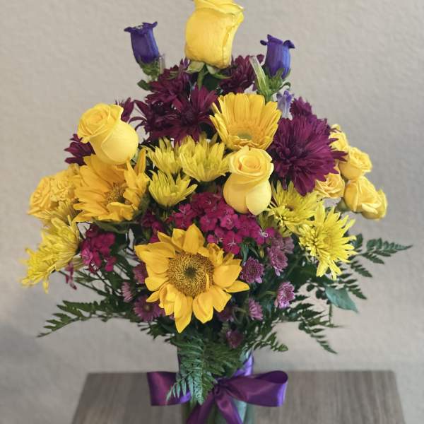 Bouquet of yellow roses, sunflowers, and purple blooms in a glass vase