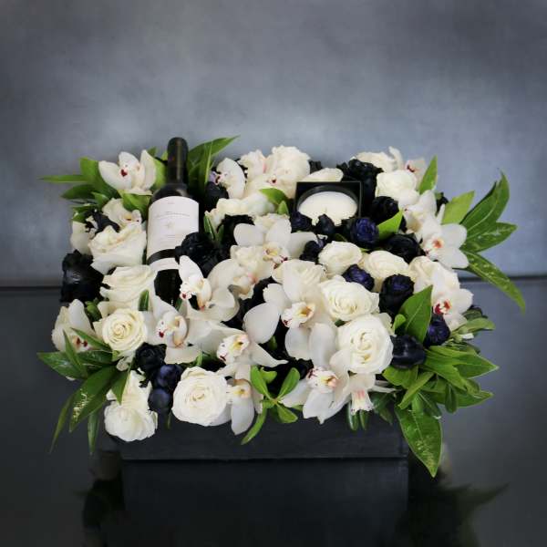 White roses and orchids arranged around a wine bottle and candle in a black box.