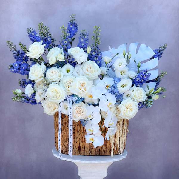 White roses and orchids arranged with blue flowers in a woven basket