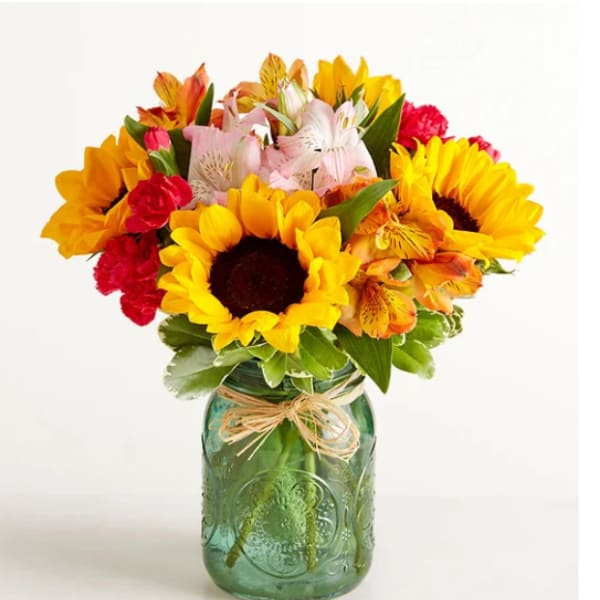 Mixed bouquet of sunflowers, alstroemeria, and carnations in a green glass jar vase
