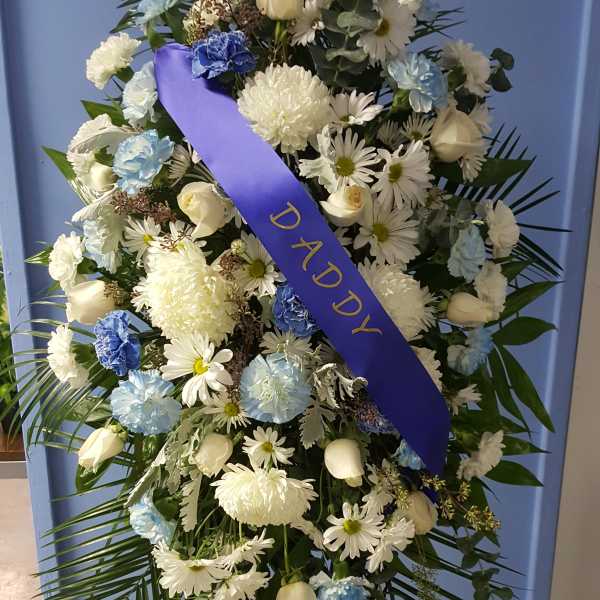 Standing funeral spray of white and blue flowers with a purple ribbon reading DADDY