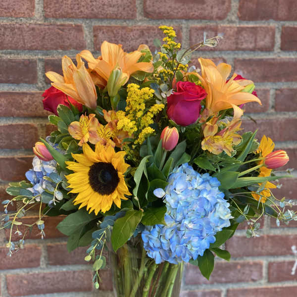 Mixed bouquet with sunflowers, roses, lilies, and blue hydrangeas in a glass vase