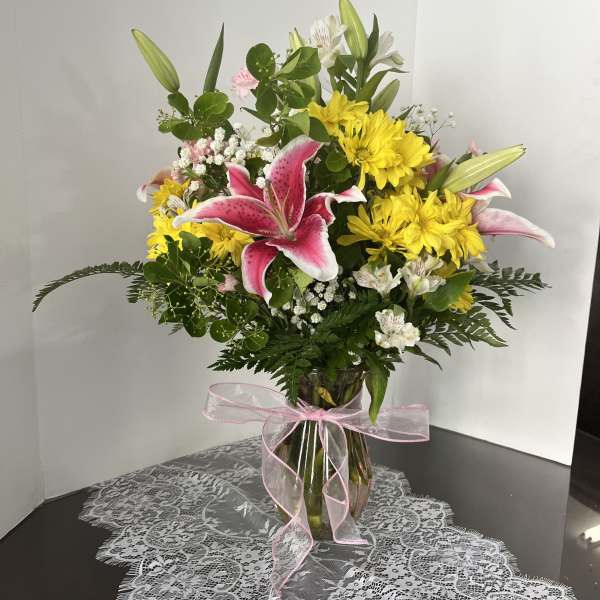 Pink lilies and yellow daisies in a glass vase with a pink ribbon