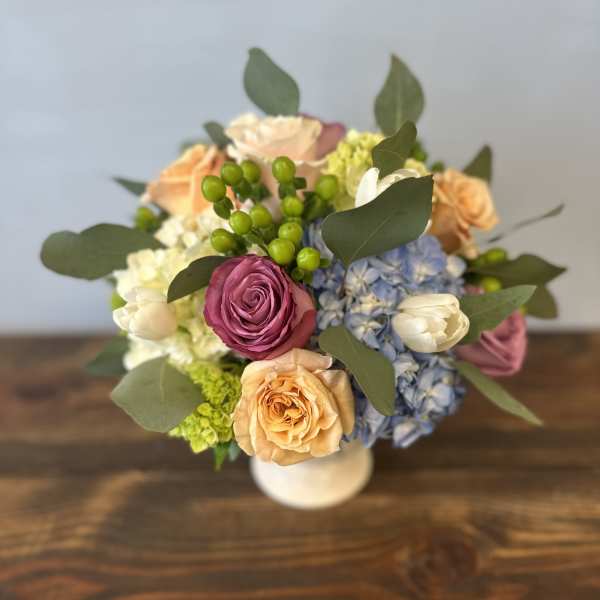 Compact arrangement of peach and mauve roses with blue hydrangeas in a white vase