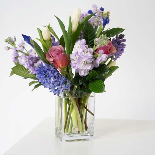 Mixed bouquet of pink roses, purple stock, blue blooms, and white tulips in a clear glass vase