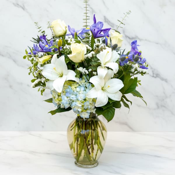 White lilies, cream roses, blue hydrangea, and purple flowers in a glass vase