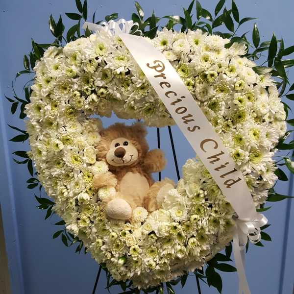 Heart-shaped white flower wreath with a teddy bear and ribbon on a stand