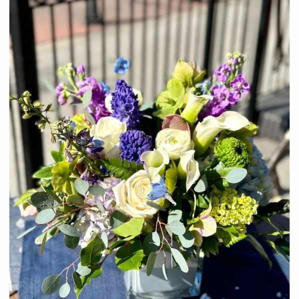 Mixed arrangement of white roses and calla lilies with purple and blue blooms in a square glass vase